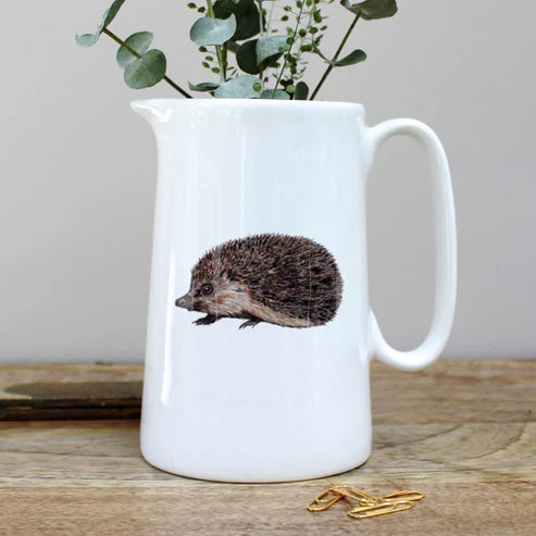 White ceramic pitcher with a hedgehog design on a wooden surface with greenery in the background