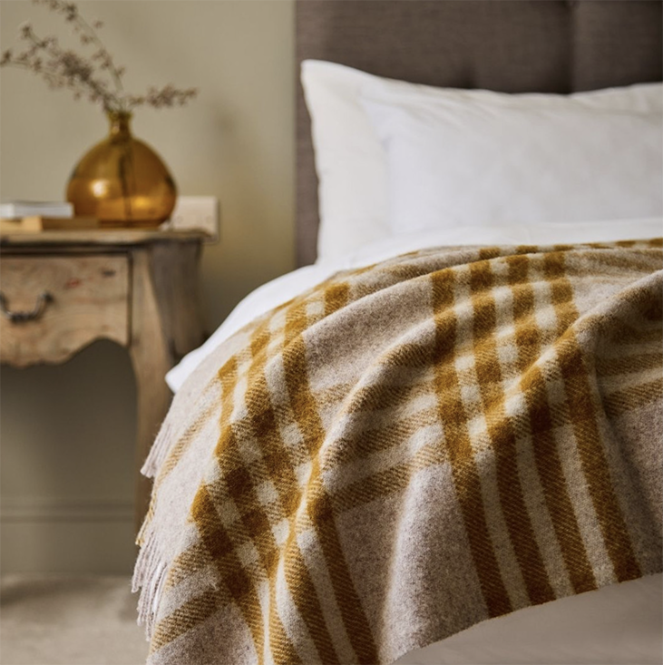 Hex English Mustard plaid blanket draped over a bed with a side table and decorative vase in the background.