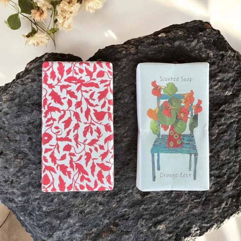Two bar of soap wrapped in paper, one with a red floral pattern and the other with a colorful chair and flowers.
