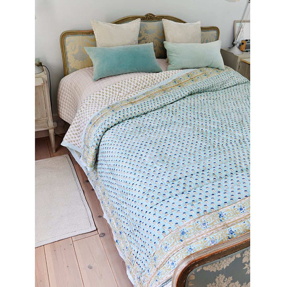 A turquoise and white block print quilt with mustard, navy, and turquoise trim, displayed on a bed in a bedroom setting.