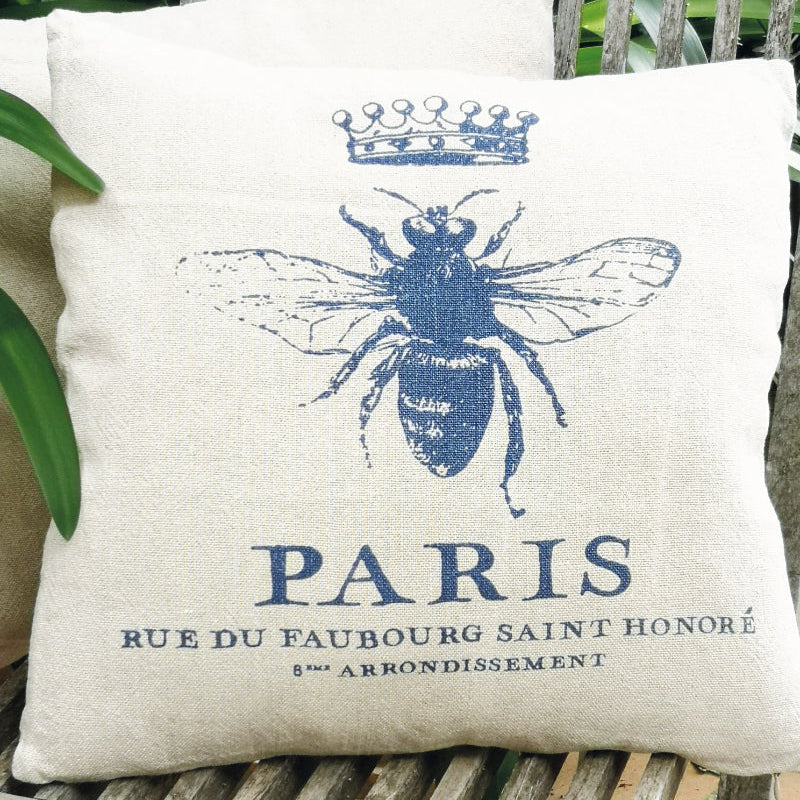 Decorative pillow with a blue bee design and 'Paris' text on a wooden bench.