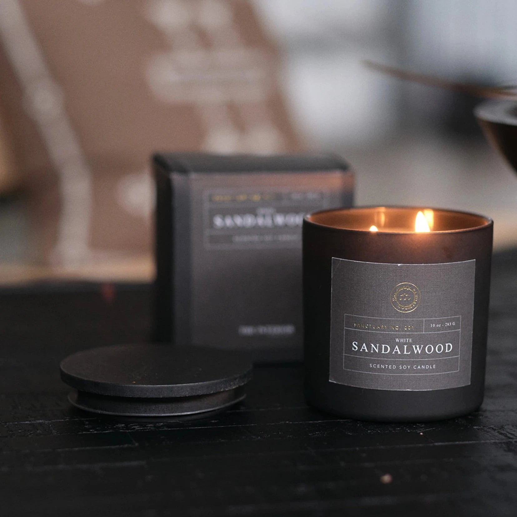 Sandalwood candle in a black container with packaging in the background on a dark surface.