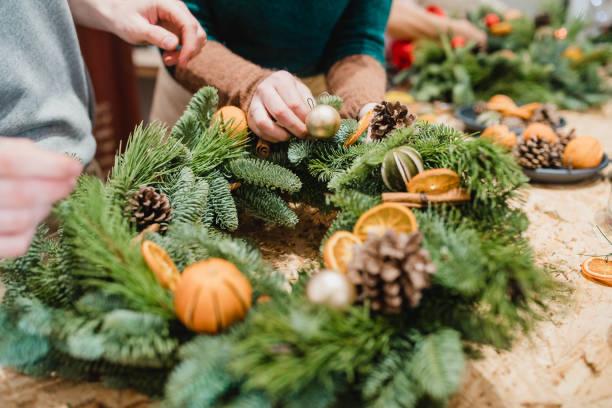 Person making a Christmas Wreath with pine cones, oranges, and other decorations on a table.