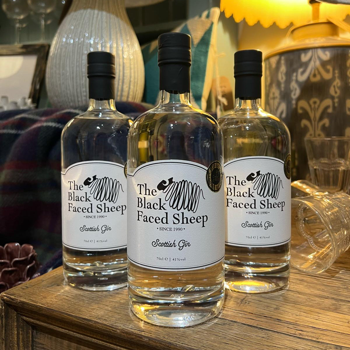 Three bottles of 'The Black Faced Sheep' gin on a wooden surface with decorative lamps in the background.