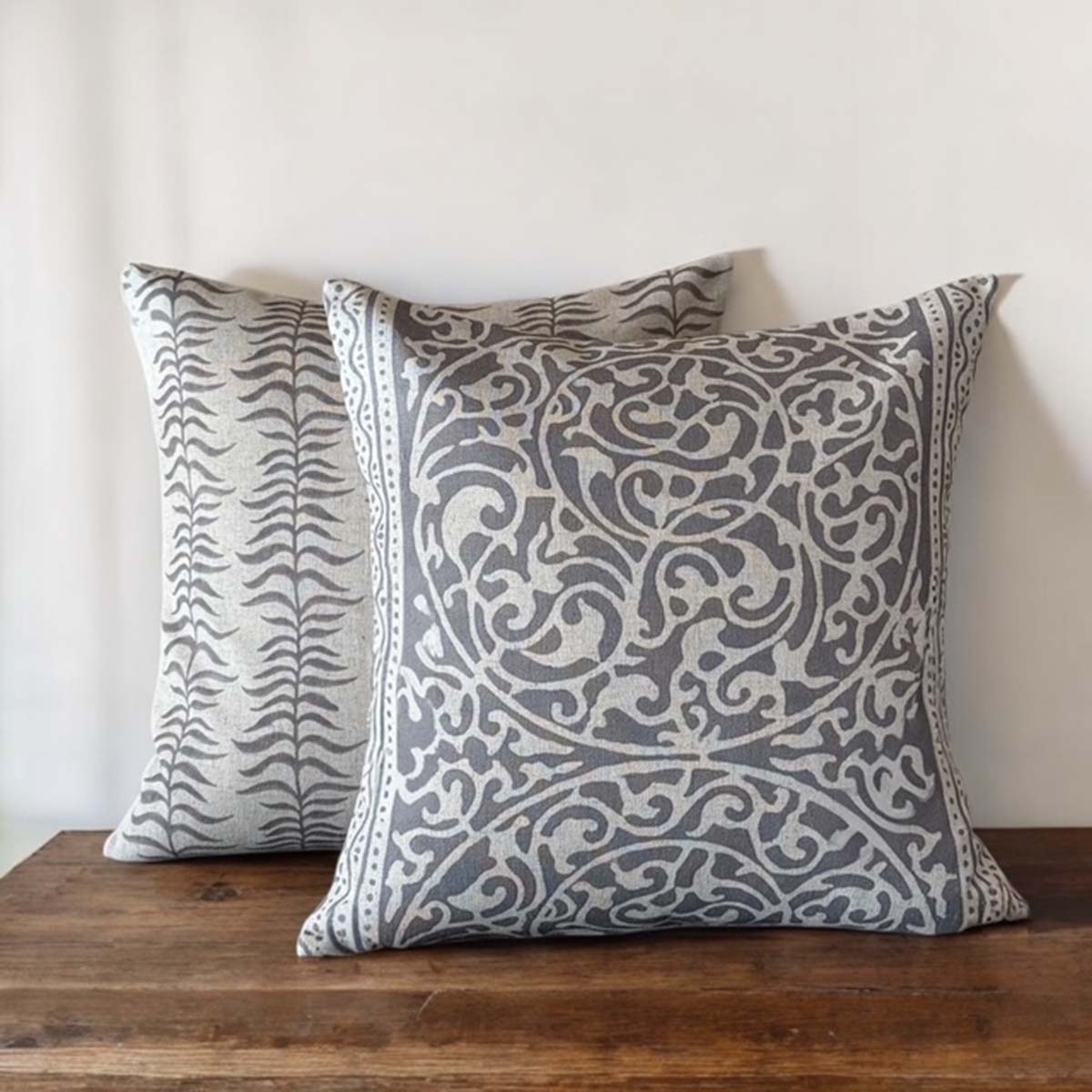 Two patterned throw cushions, the leafy stripe and fretwork designs, on a wooden surface with a neutral background.
