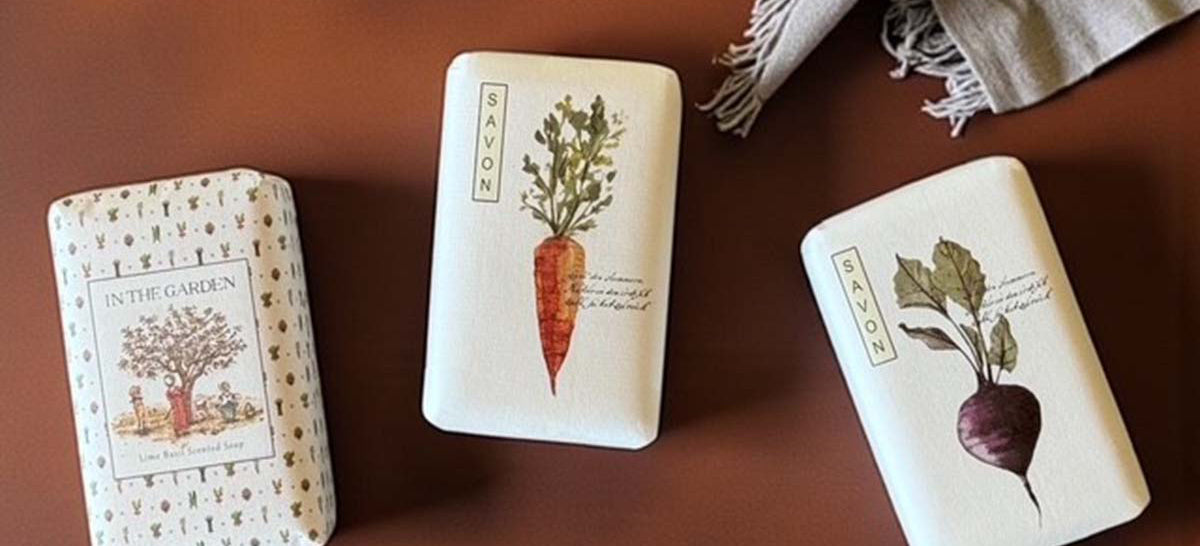 Three soap bars with vegetable illustrations on a brown surface with a candle and towel.