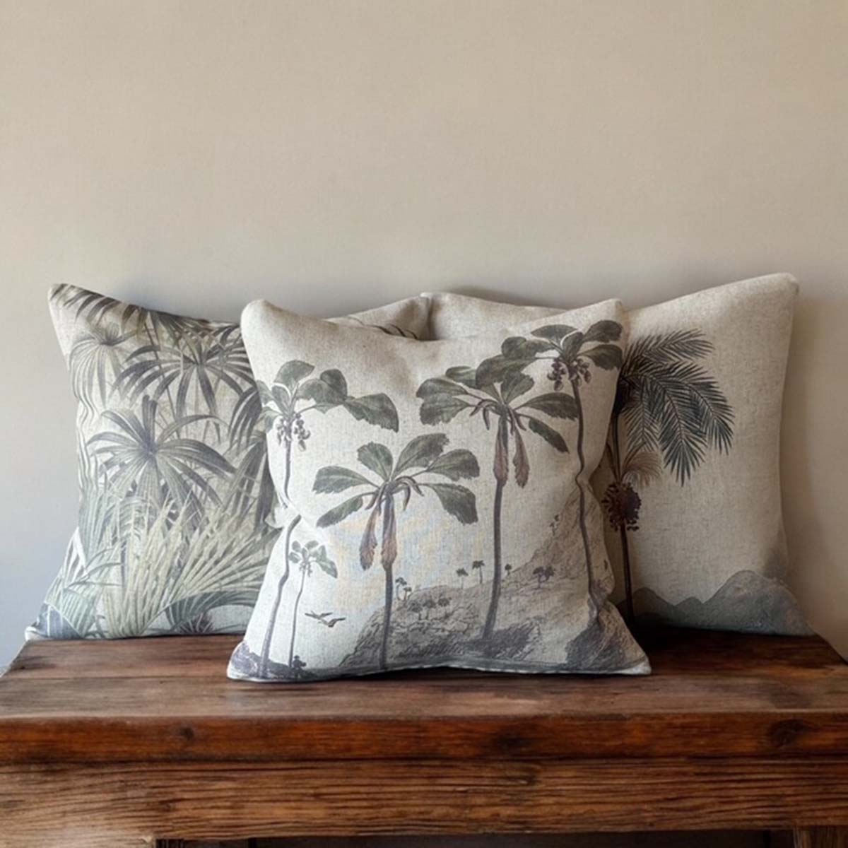 Three decorative pillows with tropical fern, tropical palm and palm tree patterns on a wooden surface against a beige wall.