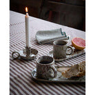 Aila porcelain mugs, a lit candle, and a half-eaten grapefruit on a striped tablecloth.