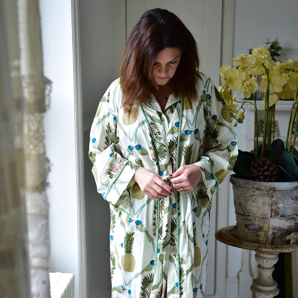 Woman wearing a floral robe standing in a room with a plant and decorative items.