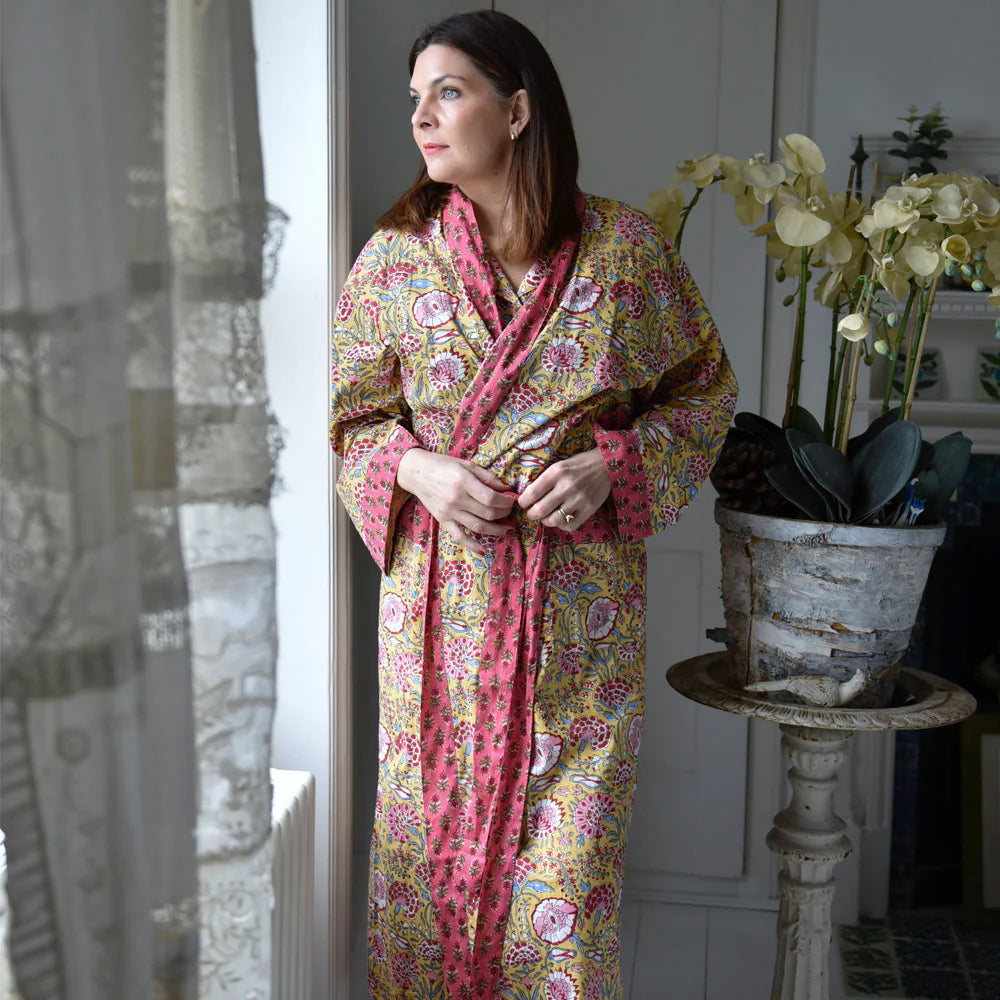 Woman wearing a colorful patterned robe standing indoors next to a window.