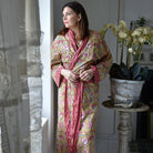 Woman wearing a colorful patterned robe standing indoors next to a window.