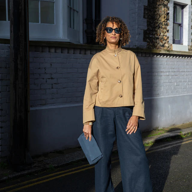 Woman in beige jacket and blue pants standing on a street with a building in the background