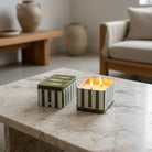 Two striped decorative candle tins with fish design on lid on a marble table with a blurred living room background.