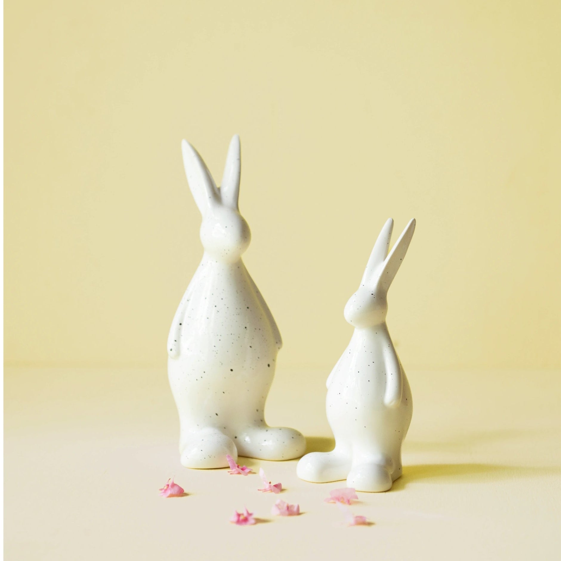 Large and small white ceramic hare figurines on a yellow background with pink flowers.
