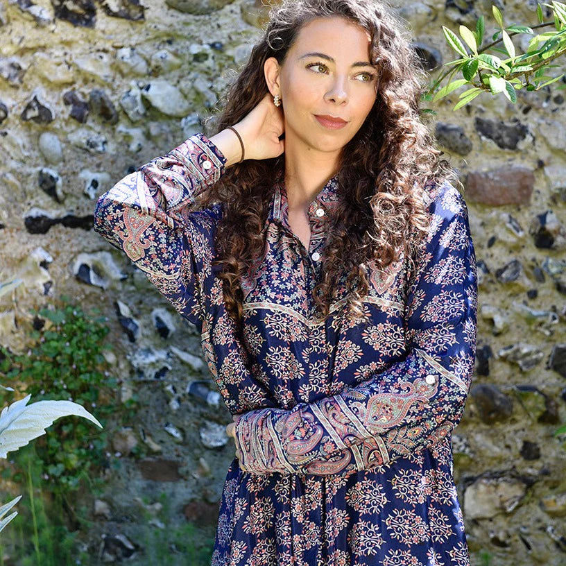 Woman wearing a patterned dress standing against a stone wall with plants.