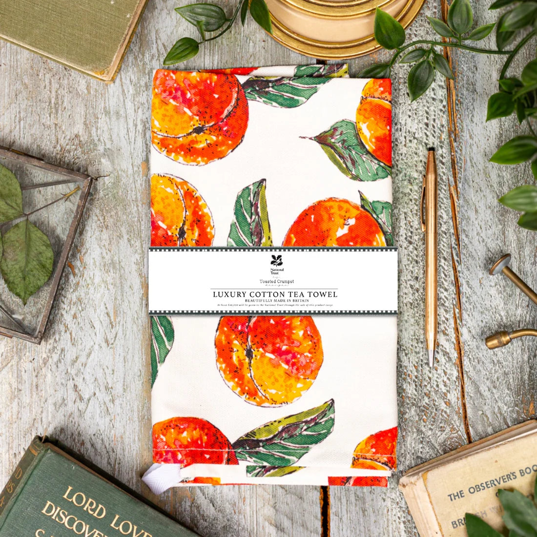 Tea towel with peach and leaf pattern on a rustic wooden surface with books and plants.