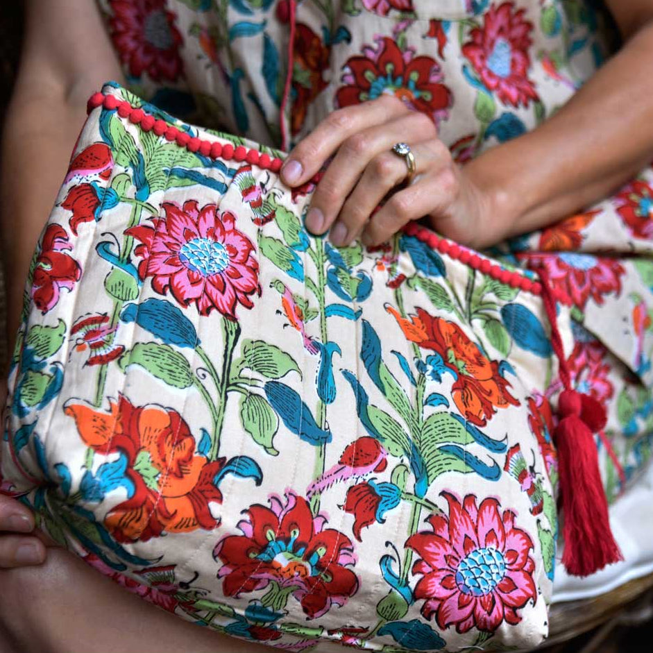 A close-up of a floral garden print wash bag with decorative piping and a pink pom-pom trim. The bag is held in someone's hand.