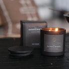 Sandalwood candle in a black container with packaging in the background on a dark surface.