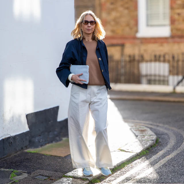 Person walking on a street wearing a navy jacket, white pants, and sunglasses.