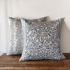 Two stone and grey patterned  cushions, the Fretwork and Leaf Stripe, on a wooden surface with a neutral background.