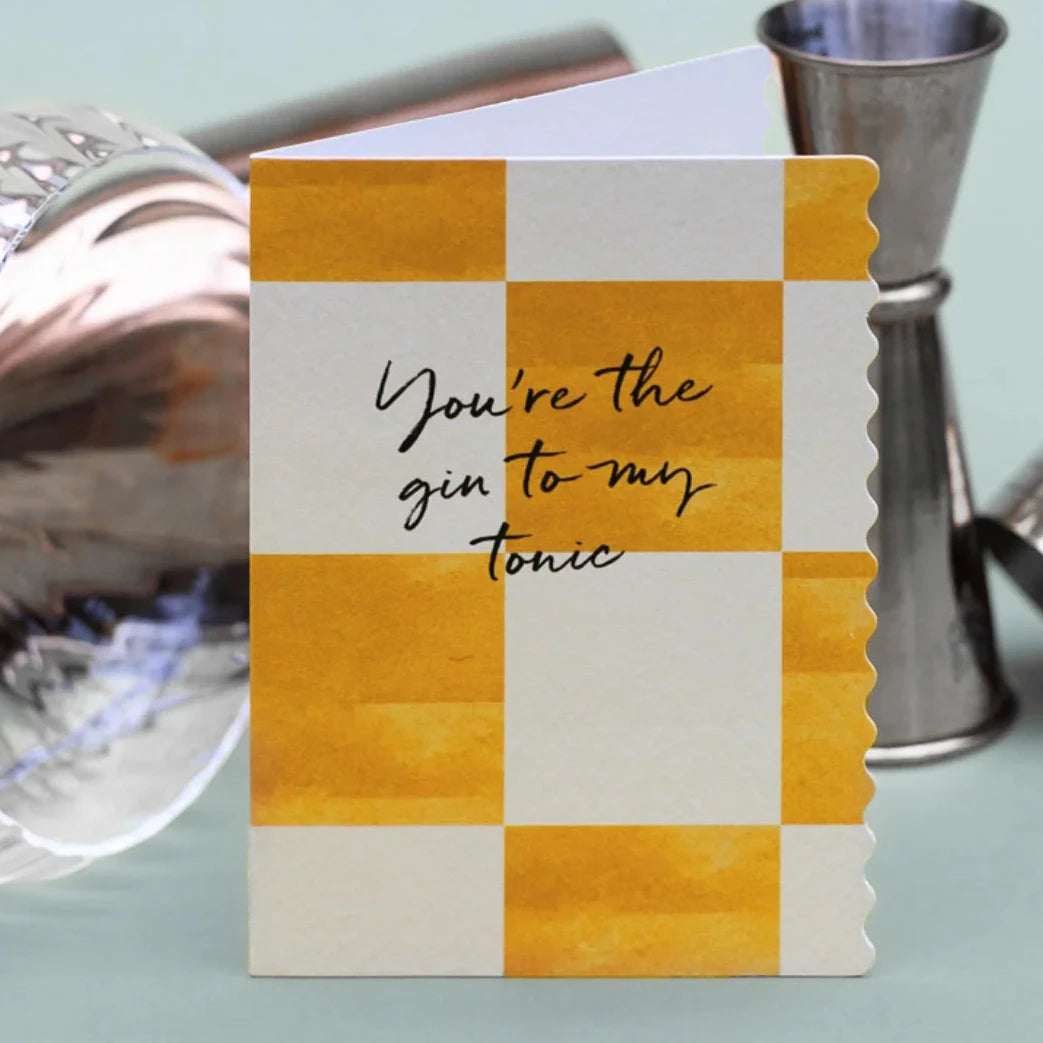 Greeting card with checkered design and text 'You're the gin to my tonic' next to cocktail shakers on a light blue background