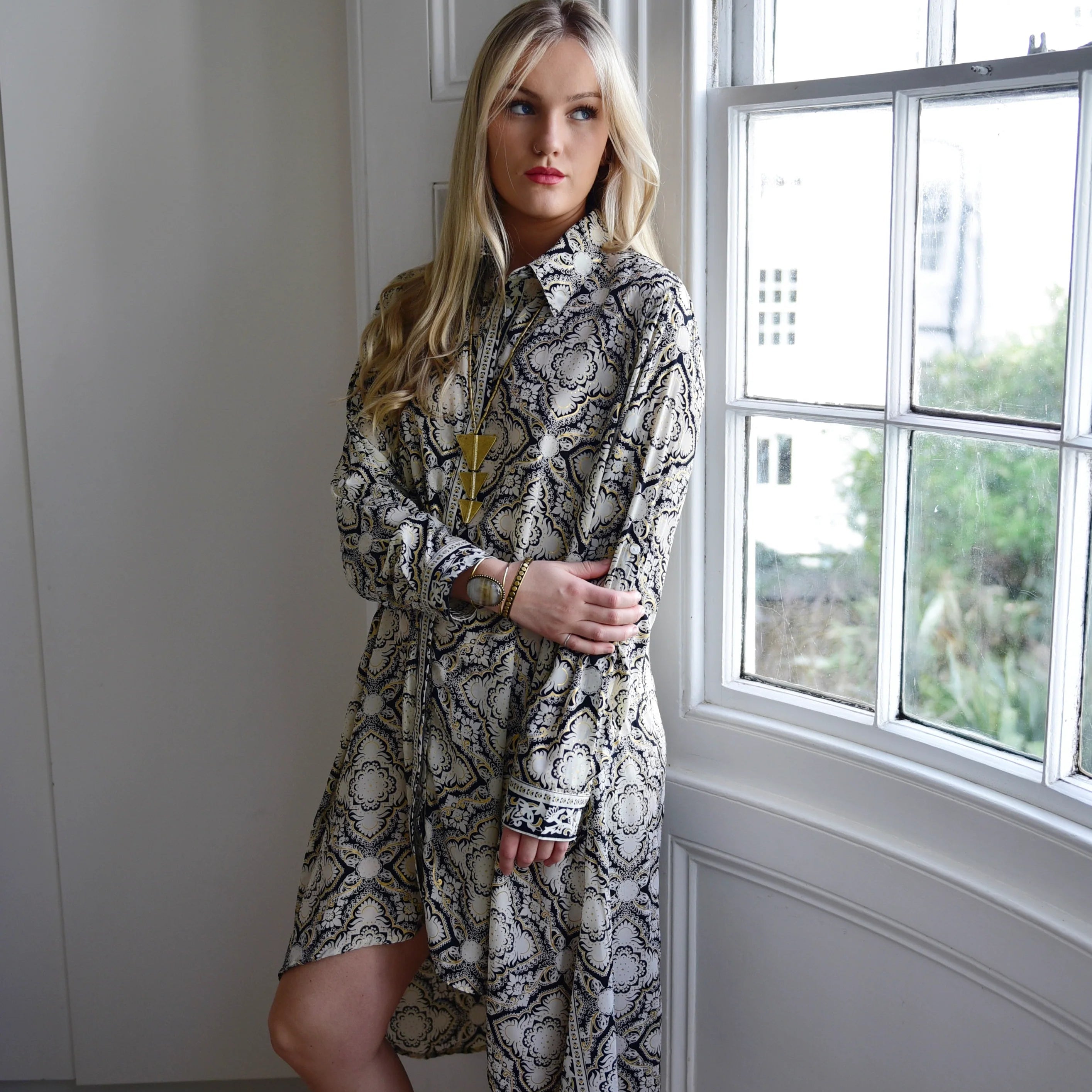 Woman in a patterned dress standing by a window with a view of buildings.